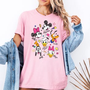 May include: Pink t-shirt with a graphic print of Mickey Mouse, Minnie Mouse, Donald Duck, Goofy, and Pluto. The characters are drawn in a cartoon style with colorful accents. The shirt has a relaxed fit and a crew neck.