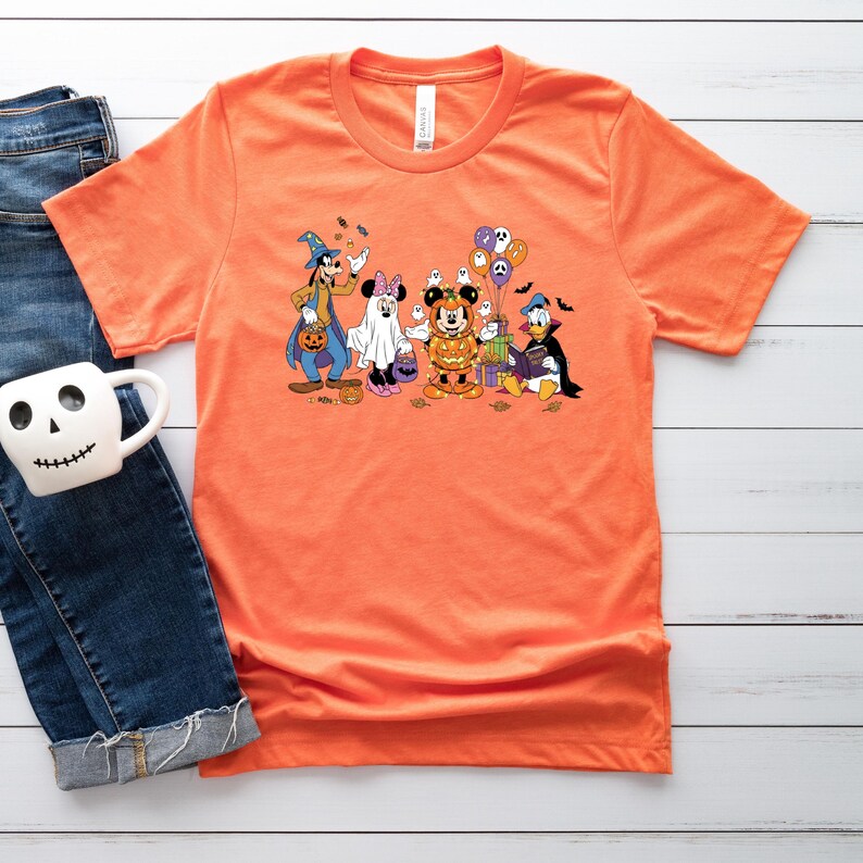 May include: Orange t-shirt featuring a Halloween-themed graphic. The graphic includes cartoon characters dressed in costumes, such as a witch, ghost, and vampire. The shirt is made of a soft material and has a crew neck.