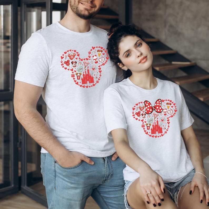 May include: A couple wearing white t-shirts with a red Mickey Mouse head design made up of hearts and Disney-themed icons. The woman's shirt features a red bow on the Mickey Mouse head.