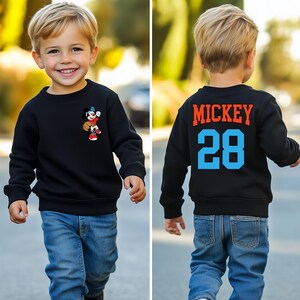 May include: Black sweatshirt with a Mickey Mouse graphic on the front and "MICKEY 28" text on the back. The child is wearing blue jeans.