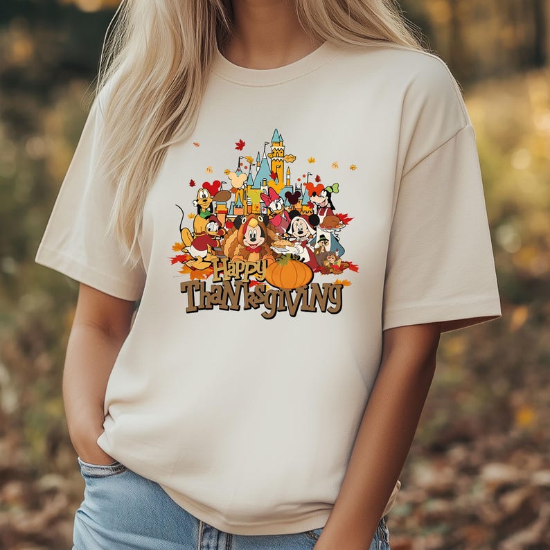May include: Beige t-shirt featuring a colorful graphic of Disney characters gathered around a pumpkin with the text "Happy Thanksgiving". The design includes a castle and autumn leaves.