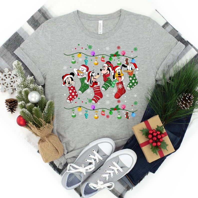 May include: A gray t-shirt with a Christmas design featuring Mickey Mouse, Donald Duck, Goofy, and Pluto wearing Christmas stockings. The characters are hanging from a string of Christmas lights.