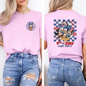 Puede incluir: Camiseta rosa claro con un dise&ntilde;o patri&oacute;tico de Pato Donald en la parte delantera y un grupo de personajes de Disney con banderas americanas en la parte trasera. La parte trasera de la camiseta tambi&eacute;n tiene el texto "4th of July Est. 1776."