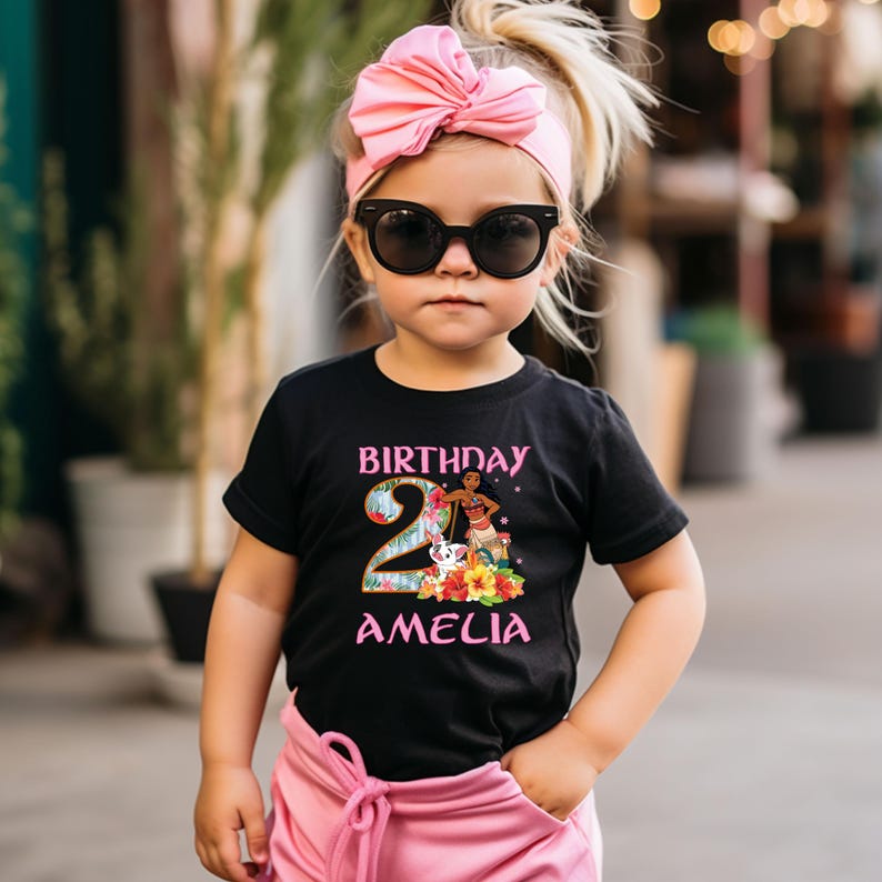 以下が含まれることがあります： ピンクとティール色の「BIRTHDAY 2 AMELIA」の文字が入った黒いTシャツ。シャツには、漫画のキャラクター、花、犬のグラフィックが描かれています。子供はピンクのリボンヘアバンドとサングラスを着用しています。