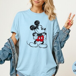May include: Light blue t-shirt featuring a black and white sketch of Mickey Mouse with red shorts. The shirt has a relaxed fit and is made of a soft material. The text "Comfort Colors Chambray" is visible in the lower right corner.