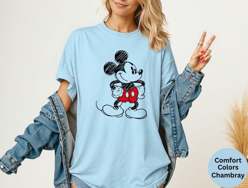 May include: Light blue t-shirt featuring a black and white sketch of Mickey Mouse with red shorts. The shirt has a relaxed fit and is made of a soft material. The text "Comfort Colors Chambray" is visible in the lower right corner.