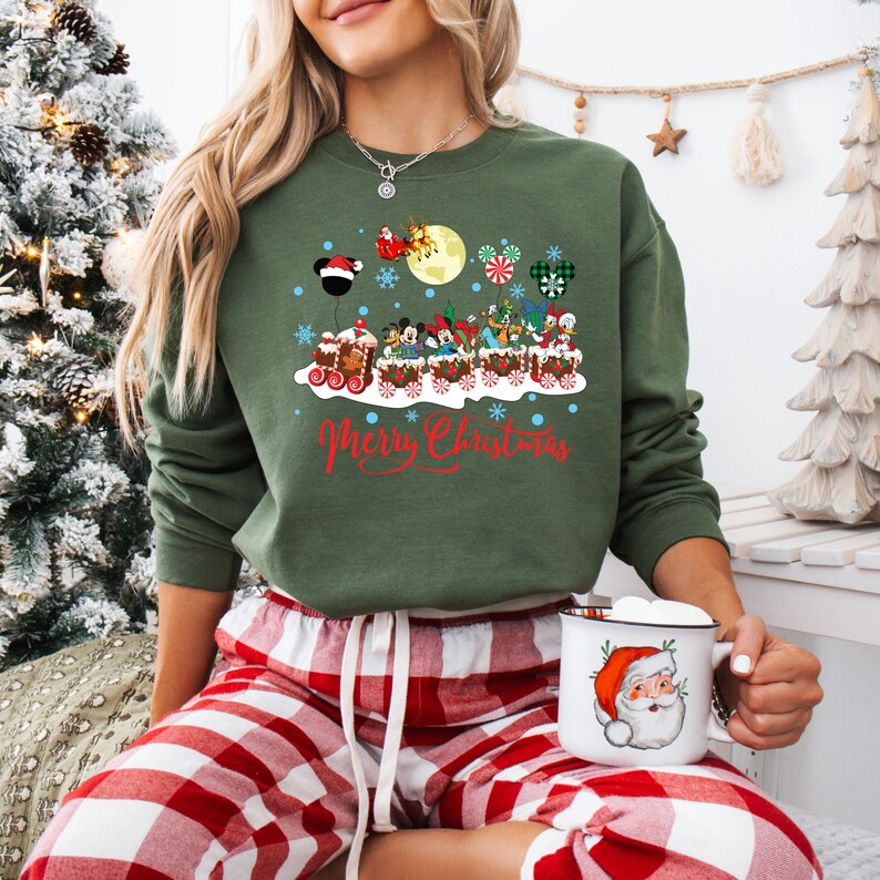 May include: A green sweatshirt with a Christmas-themed design featuring Mickey Mouse and other Disney characters riding a train. The text "Merry Christmas" is printed below the train. The model is wearing red and white plaid pajama pants and holding a white mug with a Santa Claus design.