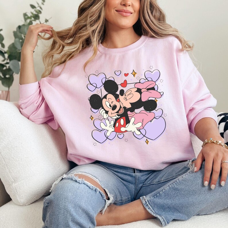May include: A pink sweatshirt with a graphic of Mickey Mouse and Minnie Mouse kissing surrounded by purple hearts. The graphic also includes small red hearts and gold stars.