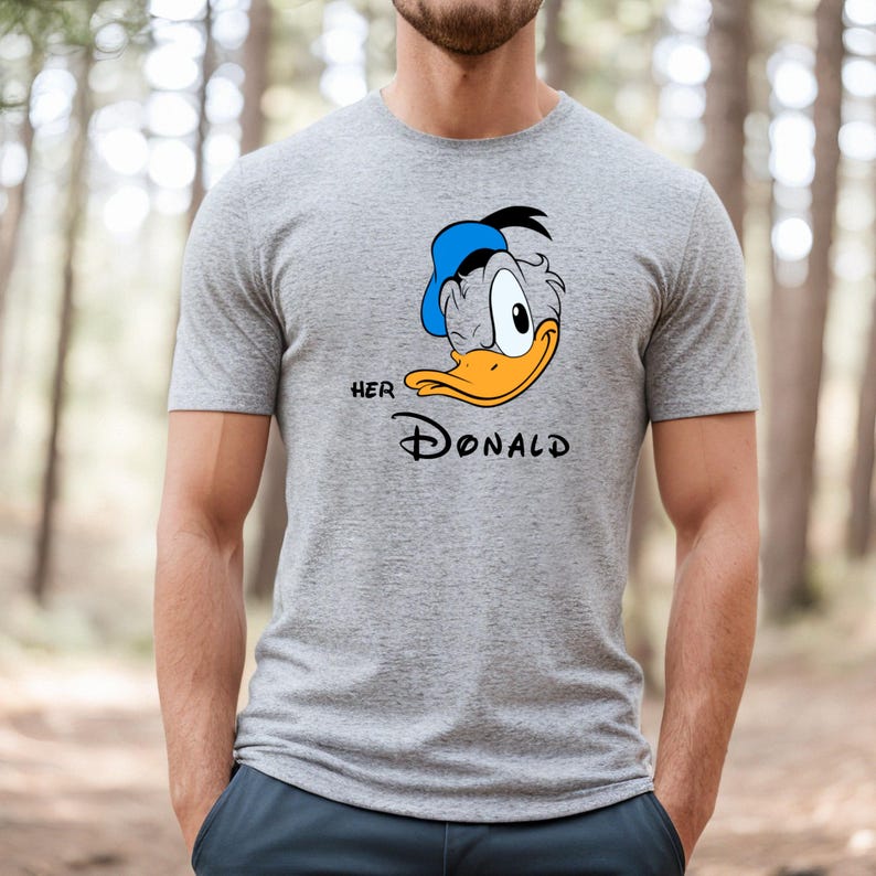 May include: A gray t-shirt featuring a cartoon image of Donald Duck wearing a blue hat. The text "HER DONALD" is printed below the image in black. The shirt is made of a soft material.