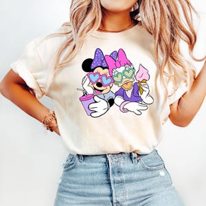 Pode incluir: Uma camiseta branca com um gr&aacute;fico de Minnie Mouse e Daisy Duck usando &oacute;culos de sol em forma de cora&ccedil;&atilde;o. Minnie est&aacute; segurando uma bebida rosa e Daisy est&aacute; segurando um cone de sorvete.