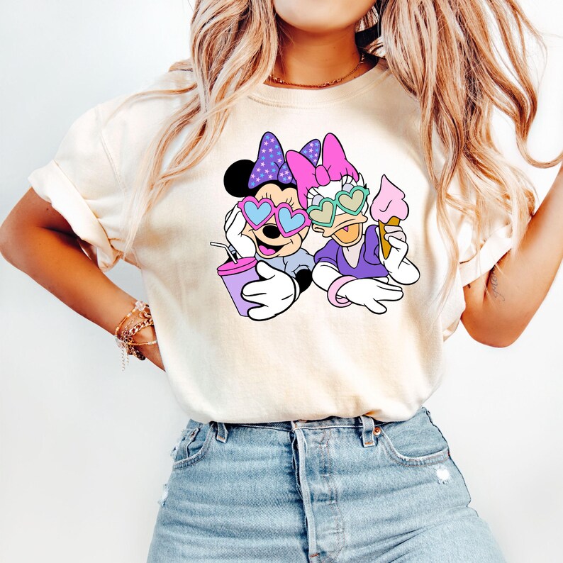 Pode incluir: Uma camiseta branca com um gr&aacute;fico de Minnie Mouse e Daisy Duck usando &oacute;culos de sol em forma de cora&ccedil;&atilde;o. Minnie est&aacute; segurando uma bebida rosa e Daisy est&aacute; segurando um cone de sorvete.
