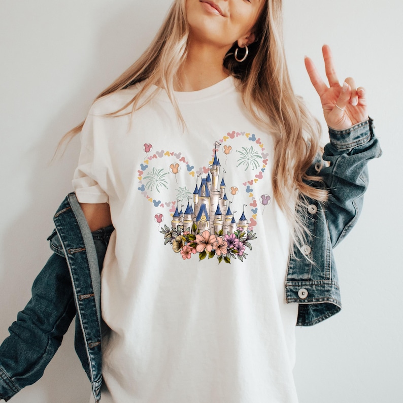 May include: Cream-colored t-shirt featuring a castle design with blue turrets, surrounded by a floral arrangement and a Mickey Mouse-shaped design with fireworks and butterflies. The shirt is a casual wear item.