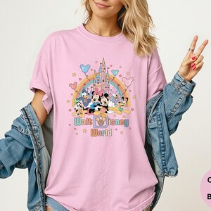 Puede incluir: Camiseta rosa claro con un gr&aacute;fico colorido de personajes de Disney, un arco&iacute;ris y un castillo. El texto "Walt Disney World" est&aacute; debajo. La camiseta es parte de la colecci&oacute;n Comfort Colors Blossom.