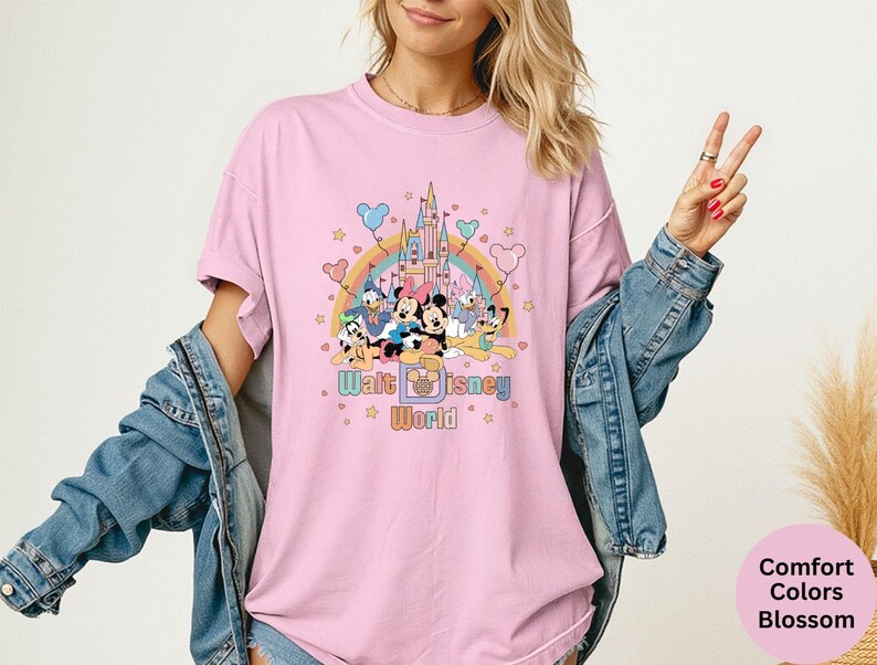 Puede incluir: Camiseta rosa claro con un gr&aacute;fico colorido de personajes de Disney, un arco&iacute;ris y un castillo. El texto "Walt Disney World" est&aacute; debajo. La camiseta es parte de la colecci&oacute;n Comfort Colors Blossom.