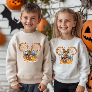 Pode incluir: Duas crian&ccedil;as vestindo moletons creme com um design de Halloween da Disney. O design apresenta um castelo, orelhas do Mickey Mouse e personagens de desenhos animados. Ab&oacute;boras e uma silhueta de morcego est&atilde;o no fundo.