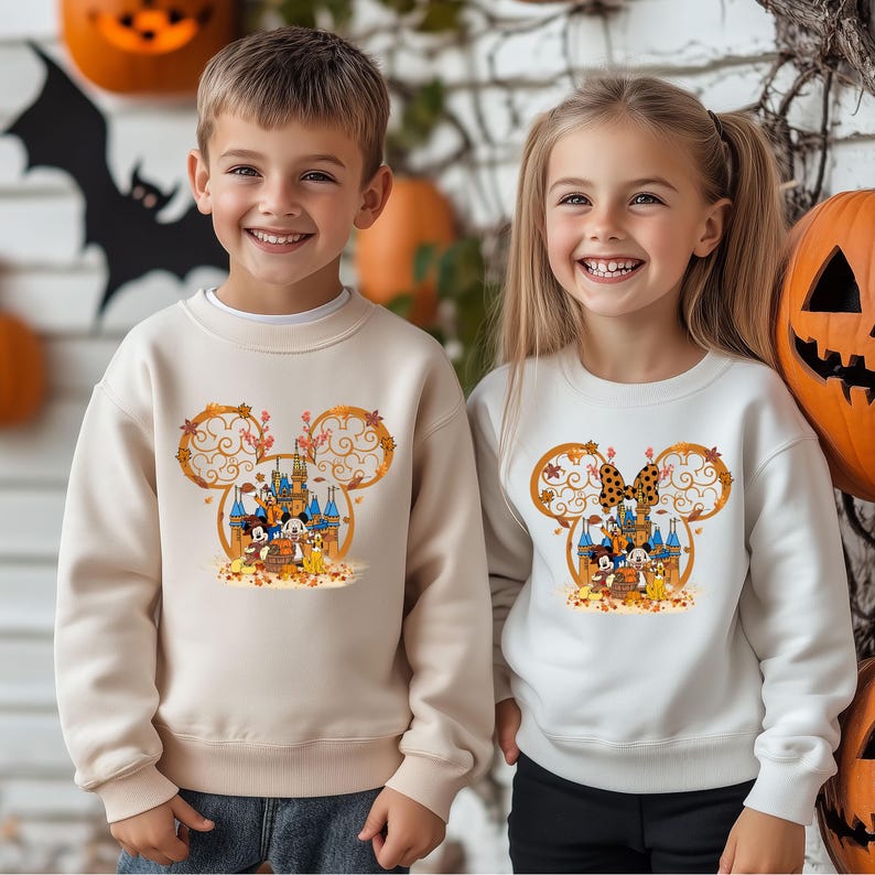 Pode incluir: Duas crian&ccedil;as vestindo moletons creme com um design de Halloween da Disney. O design apresenta um castelo, orelhas do Mickey Mouse e personagens de desenhos animados. Ab&oacute;boras e uma silhueta de morcego est&atilde;o no fundo.