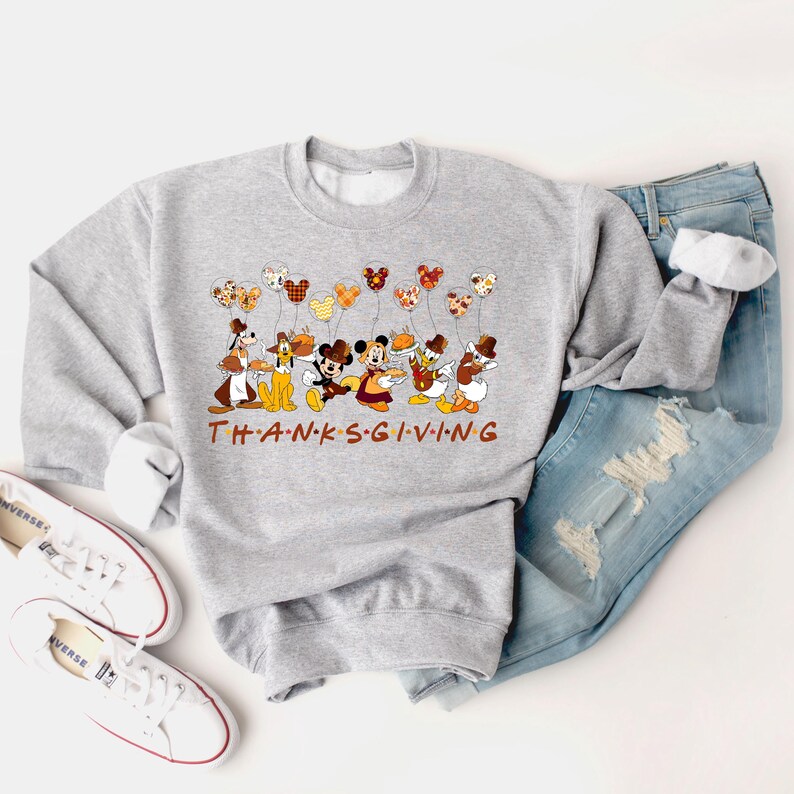 Pu&ograve; includere: Una felpa grigio chiaro con personaggi Disney che tengono in mano palloncini a tema Thanksgiving. La parola "THANKSGIVING" &egrave; stampata in lettere marroni. La felpa &egrave; abbinata a jeans blu e scarpe da ginnastica bianche.