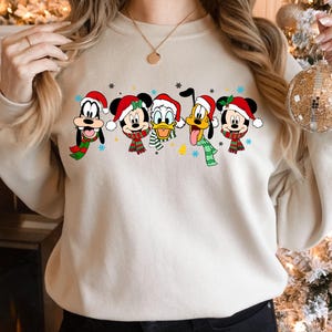 May include: A cream-colored sweatshirt with a festive design featuring Mickey Mouse, Minnie Mouse, Donald Duck, Goofy, and Pluto wearing Santa hats and scarves. The characters are arranged in a line across the front of the sweatshirt.