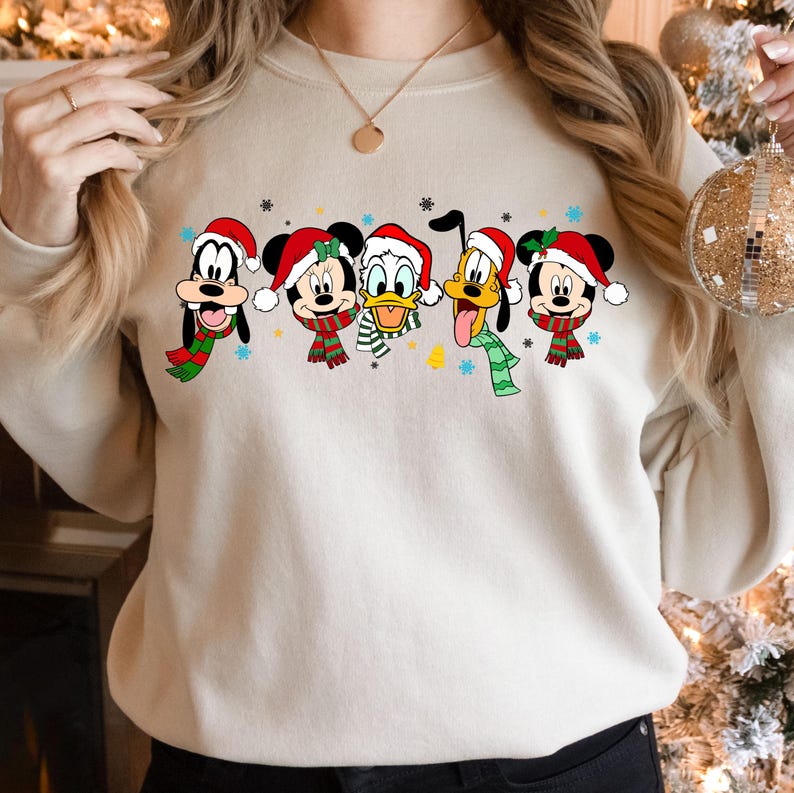 May include: A cream-colored sweatshirt with a festive design featuring Mickey Mouse, Minnie Mouse, Donald Duck, Goofy, and Pluto wearing Santa hats and scarves. The characters are arranged in a line across the front of the sweatshirt.