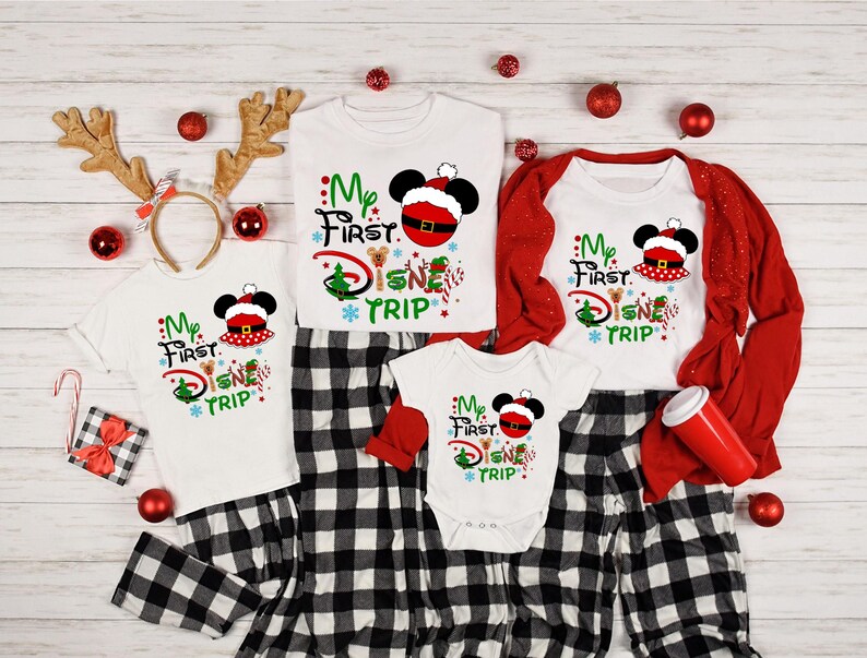 May include: A set of matching pajamas featuring a white background with a red and green Christmas-themed design. The design includes the text "My First Disney Trip" and a cartoon image of Mickey Mouse wearing a Santa hat. The pajamas are available in different sizes for adults and children.