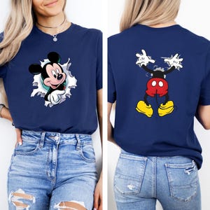 Puede incluir: Camiseta azul marino con un dise&ntilde;o de Mickey Mouse. La parte delantera muestra a Mickey Mouse saliendo de un efecto de papel rasgado. La parte trasera muestra los brazos, pantalones cortos y zapatos de Mickey. El gr&aacute;fico es en negro, rojo, amarillo y blanco.