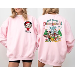 Op de afbeelding: Een lichtroze sweatshirt met een kerstthema Mickey Mouse-ontwerp op de voorkant en een Walt Disney World-afbeelding op de achterkant. Het ontwerp aan de voorkant bevat het woord "Mommy" onder de Mickey Mouse-afbeelding. De afbeelding op de achterkant bevat Mickey, Minnie, Goofy, Donald en Daisy.