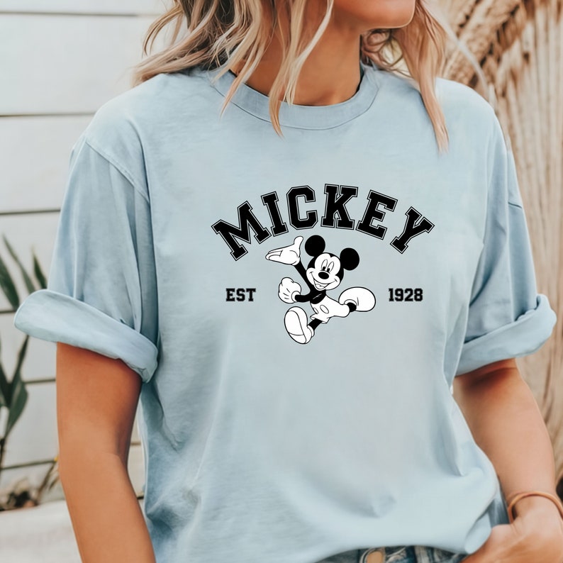 May include: A light blue t-shirt with a black and white graphic of Mickey Mouse. The graphic includes the text "Mickey" in a black arch above Mickey Mouse, "EST" to the left of Mickey Mouse, and "1928" to the right of Mickey Mouse.