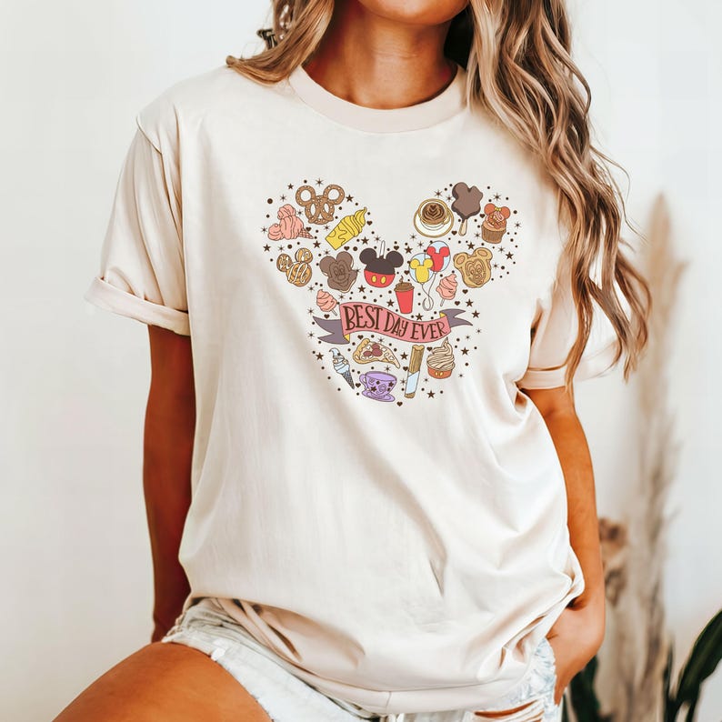 May include: A cream-colored t-shirt with a graphic of a Mickey Mouse head made up of various Disney park food items. The text "Best Day Ever" is written on a banner below the Mickey Mouse head.