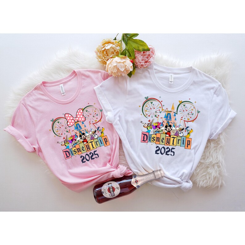 May include: Two t-shirts with a Disney Trip 2025 design. One shirt is pink with a Mickey Mouse bow and the other is white with Mickey Mouse ears. Both shirts feature a colorful illustration of Mickey Mouse and other Disney characters in front of Cinderella's Castle.