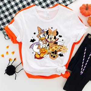 May include: White t-shirt with a graphic of Minnie Mouse and Daisy Duck dressed in Halloween costumes. Minnie Mouse is wearing a purple and orange dress with a witch hat and holding a pumpkin. Daisy Duck is wearing a witch hat and a purple and white dress. The graphic is surrounded by stars, bats, and flowers.