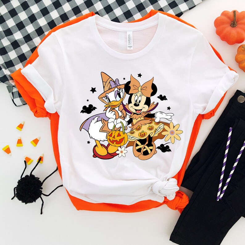 May include: White t-shirt with a graphic of Minnie Mouse and Daisy Duck dressed in Halloween costumes. Minnie Mouse is wearing a purple and orange dress with a witch hat and holding a pumpkin. Daisy Duck is wearing a witch hat and a purple and white dress. The graphic is surrounded by stars, bats, and flowers.