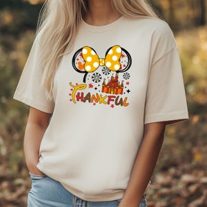 Puede incluir: Camiseta beige con un lazo amarillo de lunares y orejas negras. Debajo del lazo hay un castillo y la palabra "THANKFUL" en naranja y amarillo. La camiseta es de estilo casual.