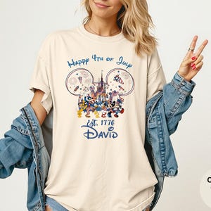 May include: Ivory-colored t-shirt with a graphic featuring Mickey Mouse and friends celebrating the 4th of July. The design includes fireworks, a castle, and the text "Happy 4th of July" and "Est. 1776 David."