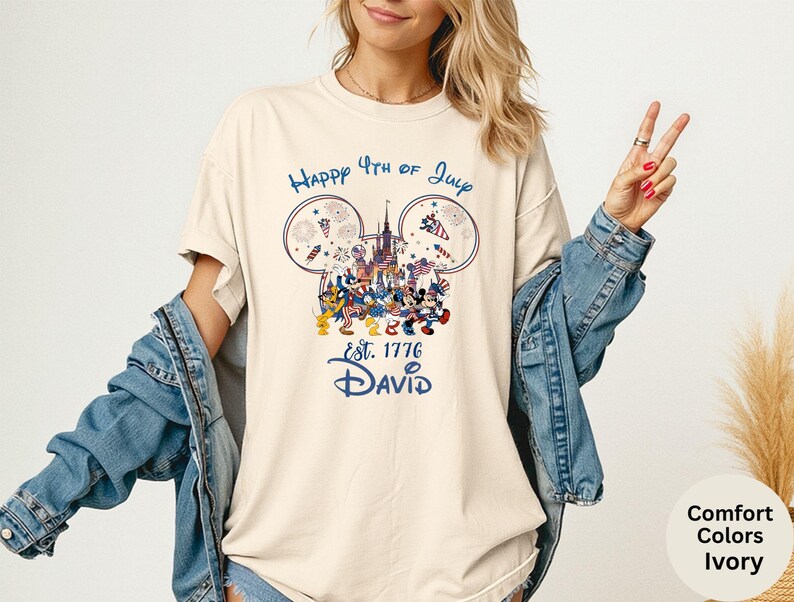 May include: Ivory-colored t-shirt with a graphic featuring Mickey Mouse and friends celebrating the 4th of July. The design includes fireworks, a castle, and the text "Happy 4th of July" and "Est. 1776 David."