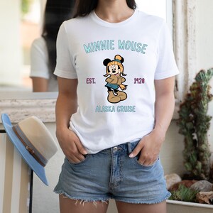 May include: White t-shirt featuring a graphic of Minnie Mouse in winter attire, with the text "MINNIE MOUSE" above the image and "ALASKA CRUISE" below. The shirt also includes "EST. 1928".