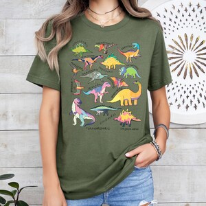 May include: A green t-shirt with a colorful dinosaur print. The dinosaurs are all different sizes and shapes, and they are arranged in a random pattern. The shirt has a crew neck and short sleeves.