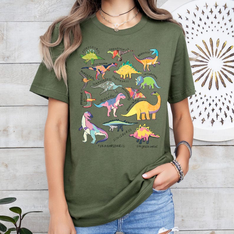 May include: A green t-shirt with a colorful dinosaur print. The dinosaurs are all different sizes and shapes, and they are arranged in a random pattern. The shirt has a crew neck and short sleeves.