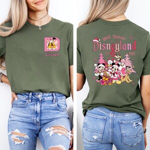 May include: Olive green t-shirt featuring a custom design on the front with a cartoon character and the word "Custom". The back of the shirt displays a Disneyland graphic with characters and text "Disneyland 1955".