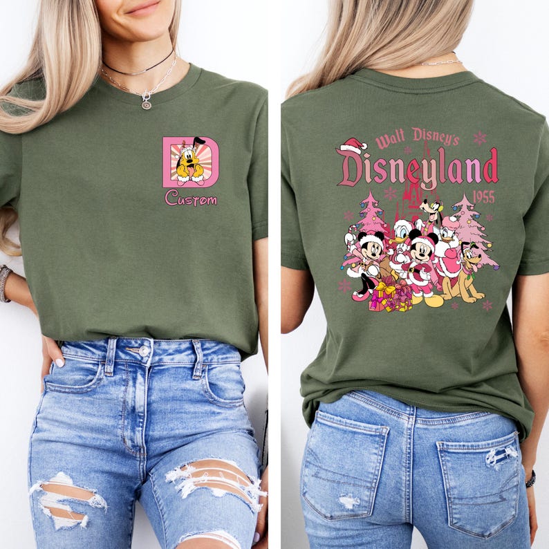 May include: Olive green t-shirt featuring a custom design on the front with a cartoon character and the word "Custom". The back of the shirt displays a Disneyland graphic with characters and text "Disneyland 1955".