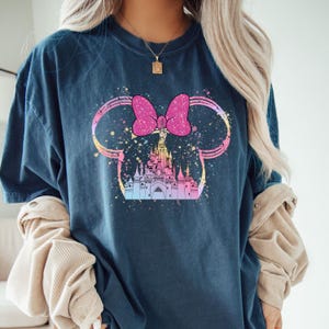 May include: A dark blue t-shirt with a colorful graphic of a castle inside of Mickey Mouse ears with a pink bow. The graphic has a watercolor effect.