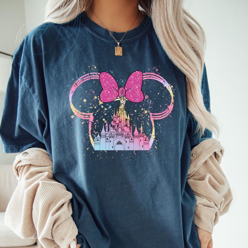 May include: A dark blue t-shirt with a colorful graphic of a castle inside of Mickey Mouse ears with a pink bow. The graphic has a watercolor effect.
