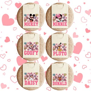 May include: Cream-colored t-shirts featuring Disney characters Mickey, Minnie, Goofy, Pluto, Daisy, and Donald. Each shirt has a character name in pink, a checkered background, and heart-shaped candies with phrases like "Love" and "Be Mine."