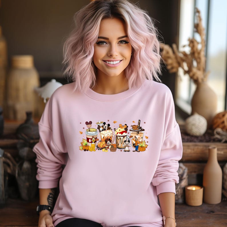 May include: A light pink sweatshirt featuring a colorful graphic of cartoon characters and fall-themed coffee drinks. The design includes pumpkins, leaves, and various characters. The sweatshirt has long sleeves and a crew neck.