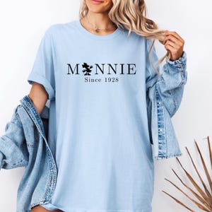 May include: Light blue t-shirt with the word "Minnie" in black, featuring a silhouette of a character and the text "Since 1928". The shirt has rolled-up sleeves and is worn with a denim jacket.