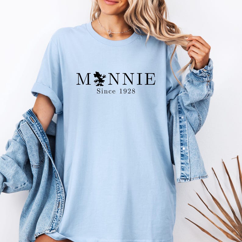 May include: Light blue t-shirt with the word "Minnie" in black, featuring a silhouette of a character and the text "Since 1928". The shirt has rolled-up sleeves and is worn with a denim jacket.