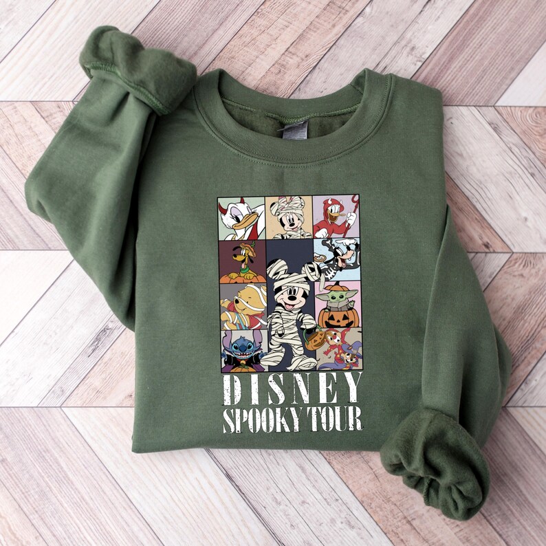 Pu&ograve; includere: Una felpa verde oliva con una grafica di personaggi Disney in costume di Halloween. La grafica &egrave; divisa in nove quadrati e include personaggi come Topolino mummia e Baby Yoda. Il testo "DISNEY SPOOKY TOUR" &egrave; stampato sotto.
