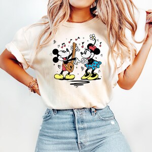 Pode incluir: Uma camiseta branca com um gr&aacute;fico de Mickey Mouse e Minnie Mouse tocando m&uacute;sica. Mickey est&aacute; tocando guitarra e Minnie est&aacute; cantando. O gr&aacute;fico &eacute; em preto, vermelho, amarelo e azul.