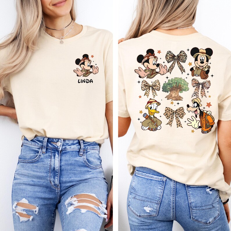 May include: A beige t-shirt with a Mickey Mouse character wearing a safari hat and holding a compass on the front. The back of the shirt features a design with multiple Mickey Mouse characters, a tree, and bows. The shirt has the name "LINDA" printed on the front.