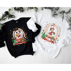Pode incluir: Camisolas pretas e brancas com um design tem&aacute;tico de Natal. O design inclui Mickey e Minnie Mouse dentro de globos de neve, rodeados por elementos festivos como flocos de neve, bengalas doces e o texto "Merry Christmas 2025".