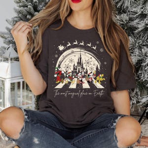 Puede incluir: Una camiseta gris oscuro con un gr&aacute;fico de Mickey Mouse y sus amigos caminando por una calle con el texto "The most magical place on Earth". El gr&aacute;fico tambi&eacute;n incluye una silueta del castillo de Cenicienta y fuegos artificiales.